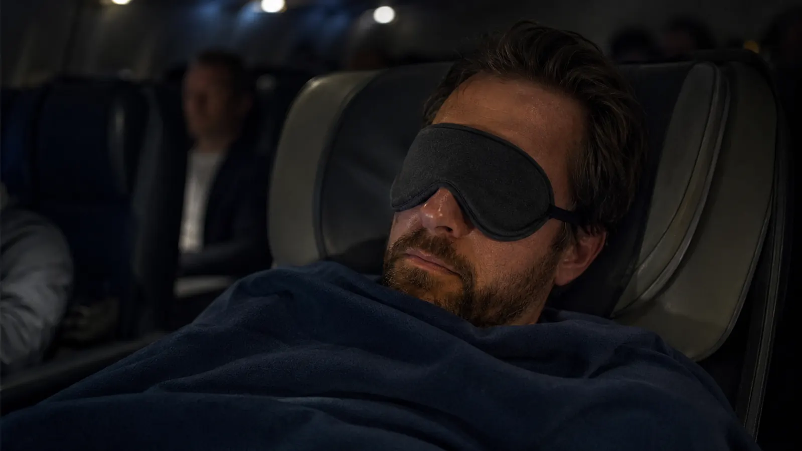 Traveler asleep on a red-eye flight wearing a contoured eye mask, blanket pulled up, cabin dark