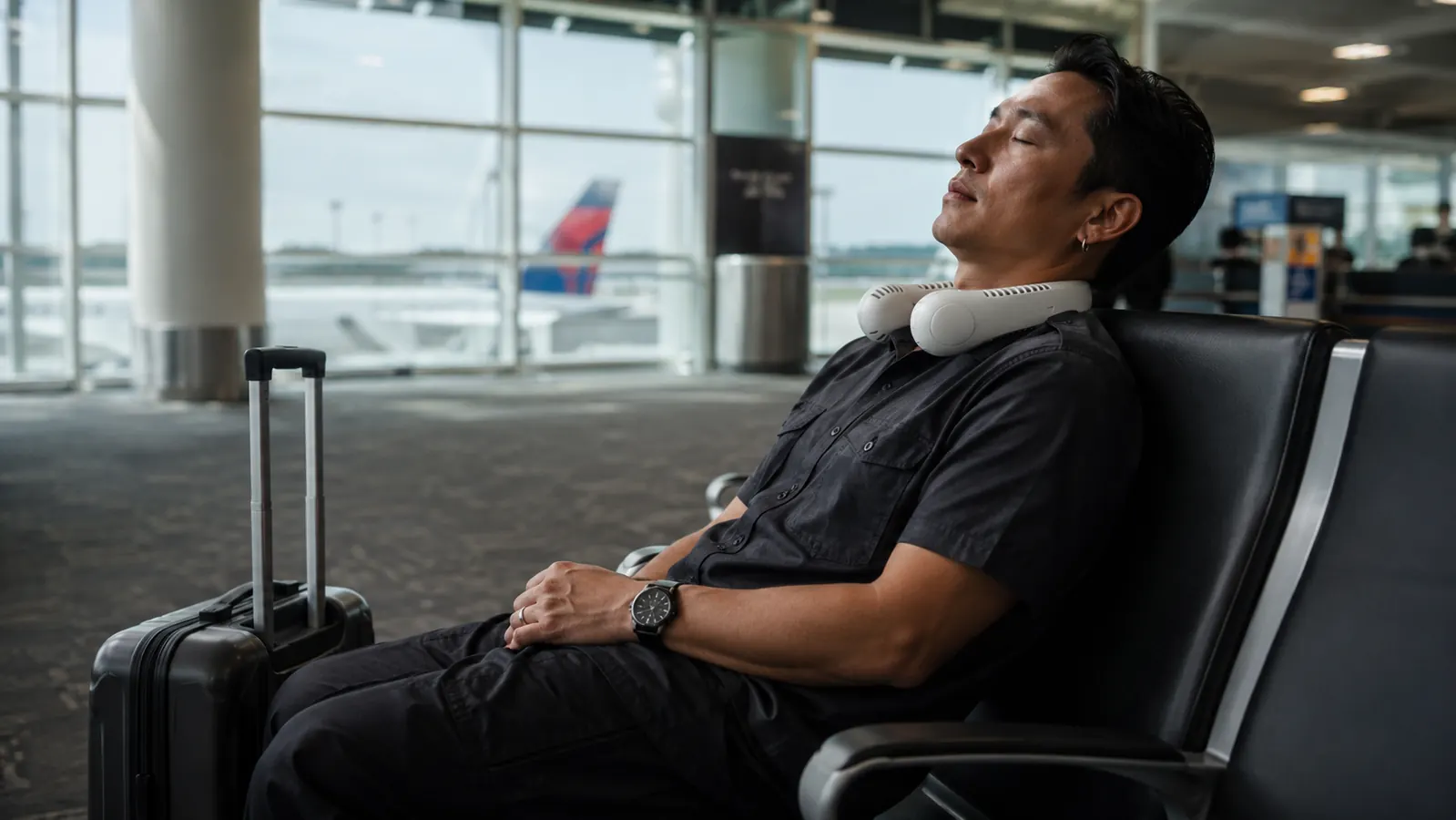 Traveler resting at an airport gate with eyes closed, neck fan still worn and running during a layover
