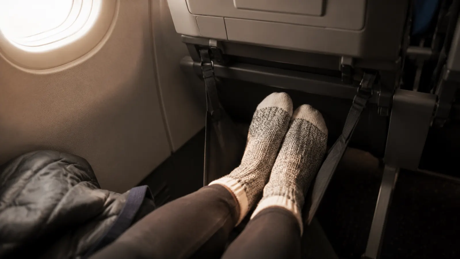 Traveler resting bare feet in an elevated fabric foot hammock under an airplane seat on a long flight