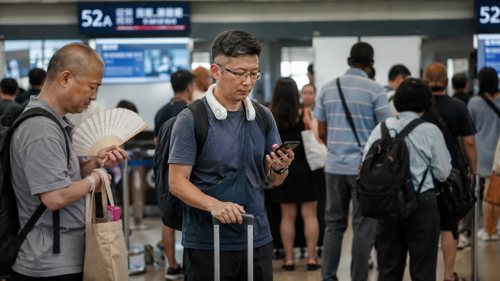 Traveler standing at a gate holding a rolling bag handle with both hands while the JISULIFE neck fan rests around their shoulders