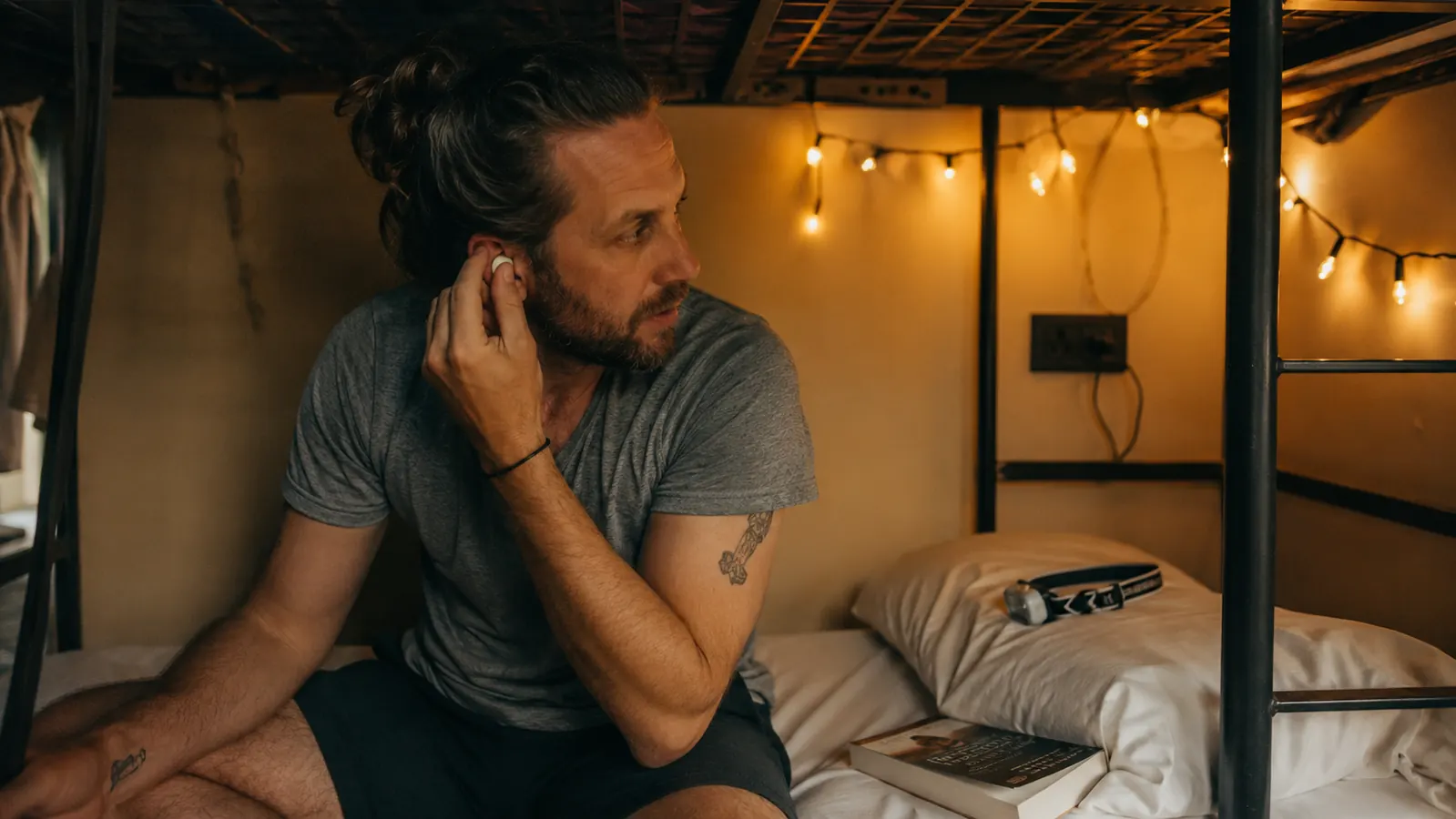 Traveler pressing a silicone earplug into place while sitting cross-legged on a hostel bunk bed with a headlamp and paperback nearby
