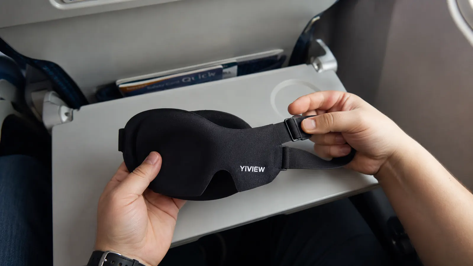 Close-up of a hand holding a black contoured 3D sleep mask with the packaging card beside it on a tray table